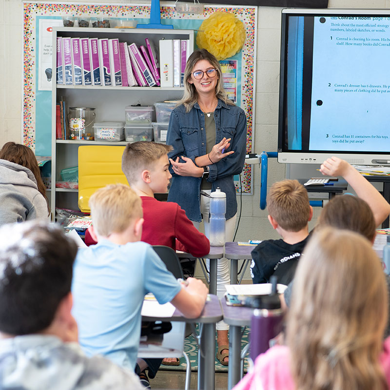 Professor in classroom with students seated on their desks.